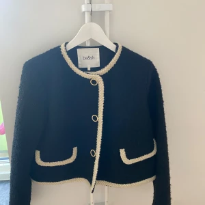 Svart jacka från ba&sh - Snygg svart jacka/kofta från det exklusiva märket  Ba&sh med kontrasterande vita detaljer. Jackan har en elegant design med guldknappar knappar framtill och en rund halsringning. Perfekt för att ge en stilren touch till din outfit. Köptes för 2199  kr och är oanvänd.