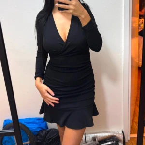 Svart omlottklänning - Snygg svart omlottklänning med v-ringning och långa ärmar. Klänningen har en tight passform och en volangdetalj vid fållen som ger en feminin touch. Perfekt för en kväll ute eller en speciell tillställning.