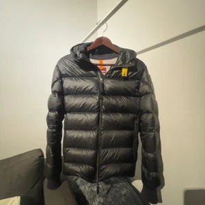 Parajumper jacka - Snygg svart dunjacka från Parajumpers med huva och dragkedja. Jackan har en broderad patch på ärmen och är perfekt för kyligare väder. Den quiltade designen ger en modern look. Skick 10/10 knappt använd.
