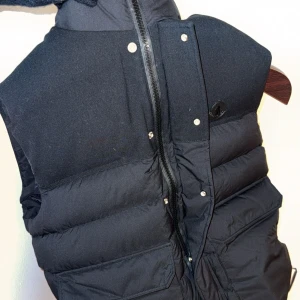 Svart dunväst från Moncler - Säljer en snygg svart dunväst från Moncler. Västen har en dragkedja och knappar framtill samt en hög krage. Perfekt för kyligare dagar och ger en stilren look. Stlig väst! Skriv för info eller vid intresse!