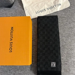 Svart halsduk från Louis Vuitton - Elegant svart halsduk från Louis Vuitton med diskret rutig design. Den har en klassisk LV-logga i ena hörnet och kommer i originalförpackning. Perfekt för att ge en lyxig touch till din outfit.
