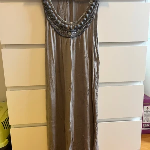 Beige långklänning från Dranella - Elegant beige långklänning från Dranella med dekorativa detaljer runt halsringningen. Klänningen har en lös passform och är perfekt för speciella tillfällen. 