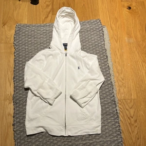 Vit hoodie från Ralph Lauren - Säljer en stilren vit hoodie från Ralph Lauren med dragkedja och det klassiska logotypbroderiet på bröstet. Perfekt för en avslappnad stil. Hoodien har långa ärmar och en bekväm passform.