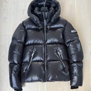 Svart kort pufferjacka från Mackage - Säljer en snygg svart pufferjacka från Mackage. Jackan har en glansig finish med dragkedja framtill och två fickor med dragkedjor. Den har en hög krage och en justerbar huva för extra värme. Perfekt för kyliga dagar! Nypris ca 10.000:-