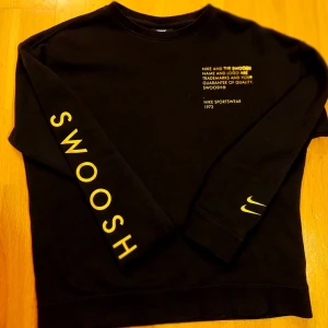 Svart tröja från Nike med guldigt text - Säljer en svart tröja från Nike med gul text 'SWOOSH' på ärmen och bröstet. Tröjan har en rund hals och långa ärmar. Perfekt för en sportig look eller avslappnad stil. 🖤💛
