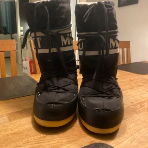 Svarta Moon Boots med snörning - Säljer ett par svarta Moon Boots med snörning och vit textdetalj. Perfekta för kalla vinterdagar med sin varma och vadderade insida. De har en robust sula i gummi som ger bra grepp på snö och is. Dem har lite slitningar på tårna och på smörerna och det är även ett slitet namn skrivet med bläck inuti som jag försökt få bort. Priset kan ändras om någon kommer med ett bra förslag. Skriv för fler bilder❤️