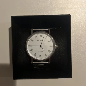 Klassisk klocka från Modiva - Snygg och stilren klocka från Modiva med vit urtavla och romerska siffror. Klockan har en silverfärgad boett och ett matchande armband. Perfekt för en elegant look.