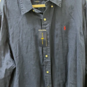 Mörkblå skjorta från Ralph Lauren - Snygg mörkblå skjorta från Ralph Lauren med klassisk krage och knappar framtill. Skjortan har en röd broderad logotyp på bröstet och är i en custom fit. Perfekt för en stilren look. Helt ny , aldrig använd. Pris kan diskuteras vid snabb affär. Nytt pris 1799kr.