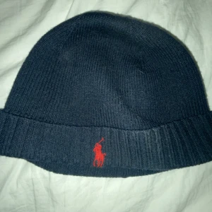 Mörkblå stickad mössa från Polo Ralph Lauren - Snygg mörkblå stickad mössa från Polo Ralph Lauren med röd broderad logotyp framtill. Perfekt för kyliga dagar med sin ribbade kant som ger extra värme och stil.