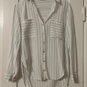 Vit  randig skjorta från Mango - Snygg vit skjorta från Mango med svarta ränder och två bröstfickor. Skjortan har långa ärmar och knappar framtill. Perfekt för en stilren look.