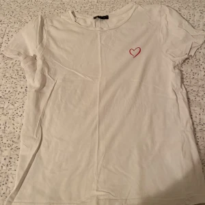 Vit t-shirt med hjärtdetalj från Amisu - Säljer en söt vit t-shirt från Amisu med ett litet rött hjärta broderat på bröstet. T-shirten har en klassisk rund hals och korta ärmar.💖 PRIS KAN DISKUTERAS