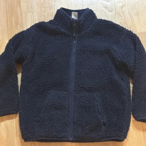 Svart teddyjacka med dragkedja - Mysig svart teddyjacka med dragkedja framtill. Jackan har en hög krage och långa ärmar, perfekt för kyliga dagar. Den fluffiga texturen ger en trendig look och den är lätt att matcha med olika outfits.