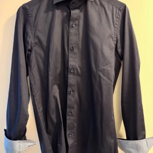 Svart skjorta med knappar - Snygg svart skjorta med långa ärmar och knappar framtill. Perfekt för en stilren look. Ärmsluten har en kontrasterande randig insida som ger en extra touch när de viks upp. Endast använd en gång.