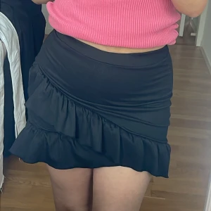 Svart kortkjol med volang - Snygg svart kortkjol med volangdetaljer. Kjolen har en tight passform och är perfekt för att ge en stilren look. Den är enkel att matcha med olika toppar och accessoarer.