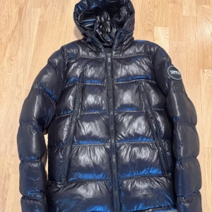 Svart pufferjacka från Supply - Snygg svart pufferjacka från Supply & demand med glansig finish och huva. Jackan har dragkedja framtill och två sidofickor med dragkedjor. Perfekt för kyliga dagar. Nypris: 3000 kr.
