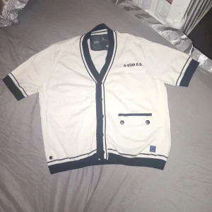 Vit kortärmad kofta från G-Star - Säljer en stilren vit kofta från G-Star med korta ärmar och svarta detaljer. Koftan har en v-ringad design och två knappar på en ficka framtill. Perfekt för en avslappnad look.