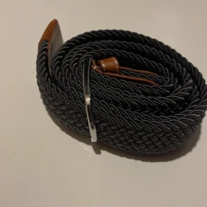  flätat bälte - Snyggt grått flätat bälte med bruna läderdetaljer och silverfärgat spänne. Perfekt för att ge en stilren touch till din outfit.