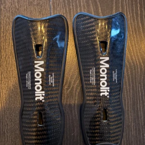 Svarta benskydd från Monolit - Säljer ett par svarta benskydd från Monolit i kolfiber. De har en ergonomisk form och är designade för att ge maximalt skydd och komfort. Perfekta för fotbollsspelare som söker högkvalitativ utrustning.
