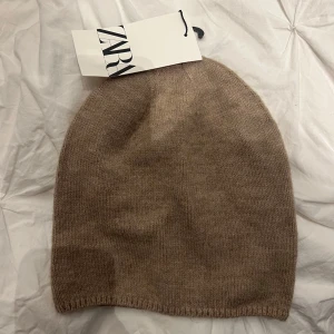 Beige stickad mössa från Zara - Säljer en stilren beige stickad mössa från Zara. Perfekt för kyliga dagar med sin mjuka och bekväma passform. Mössan är enkel och tidlös, vilket gör den lätt att matcha med olika outfits. Med lappen kvar! Köpt i NYC!
