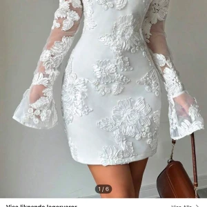 Vit klänning med blommig spets - Elegant vit klänning med blommig spetsdetalj och långa ärmar. Klänningen har en tight passform och är perfekt för speciella tillfällen. Den blommiga spetsen ger en romantisk touch och de genomskinliga ärmarna adderar en extra dimension. Aldrig använd, storlek S