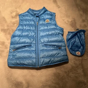 Blå dunväst från Moncler - Säljer en snygg blå dunväst från Moncler. Västen har en dragkedja framtill och två praktiska fickor med dragkedjor. Perfekt för kyligare dagar och kommer med en matchande påse för enkel förvaring.