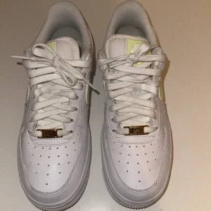 Nike Air Force i vitt och lime - Snygga Nike Air Force sneakers i vitt med limegröna detaljer. Skorna har klassisk snörning och perforeringar på tån för ventilation. Den ikoniska Nike-loggan syns på sidan och hälen. Perfekta för en stilren look.