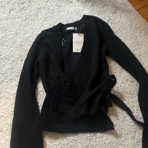 Svart omlottkofta från Gina tricot  - Säljer en elegant svart omlottkofta från Gina.Koftan har långa ärmar och ett knytband i midjan för en justerbar passform. Perfekt för lager-på-lager under kyliga dagar. 🖤 Aldrig andvänd 