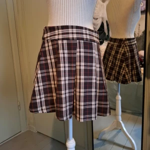 Rutig kjol från FB Sister - Snygg rutig kjol från FB Sister i brunt, vitt och rött. Kjolen har en dragkedja på sidan och ett klassiskt rutmönster som ger en stilren look. Perfekt för att skapa en trendig outfit.