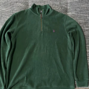 Grön tröja med dragkedja från Ralph Lauren - Säljer en stilren grön tröja från Ralph Lauren med halv dragkedja och broderad logga på bröstet. Perfekt för en avslappnad och snygg look. Tröjan har långa ärmar och är tillverkad i ett mjukt material.