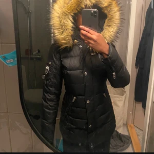 Svart dunjacka med päls från Rockandblue - Snygg svart dunjacka från Rockandblue med en stor, fluffig päls på huvan. Jackan har dragkedja och knappar framtill samt flera praktiska fickor med dragkedjor. Perfekt för kalla dagar.