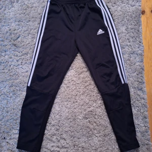 Svarta träningsbyxor från Adidas - Säljer ett par svarta träningsbyxor från Adidas med klassiska vita ränder längs sidorna. Byxorna har en dragkedja vid bensluten och elastisk midja för extra komfort. Perfekta för sport och aktivitet.