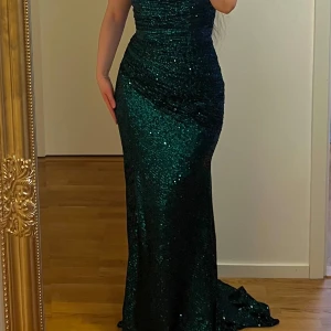 Grön glittrig långklänning - Säljer en fantastisk grön glittrig långklänning med offshoulder-design. Klänningen har en elegant slits och en figurnära passform som framhäver silhuetten. Slitsen kan sprättas upp till låret, precis som den silvriga (obs exempelklänning - exakt samma modell fast olika klänningar). Perfekt för en glamorös kväll! Använt endast en kväll. 