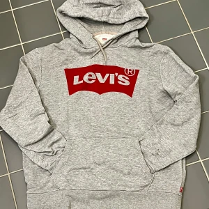 Grå hoodie från Levi's - Säljer en snygg grå hoodie från Levi's med det klassiska röda logotyptrycket på bröstet. Tröjan har en bekväm passform med långa ärmar och en praktisk huva med dragsko. 