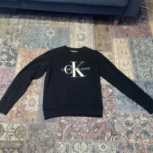 Svart tröja från Calvin Klein Jeans - Säljer en stilren svart tröja från Calvin Klein Jeans med det ikoniska CK-logotypen tryckt på framsidan. Tröjan har långa ärmar och en rund halsringning. Perfekt för en casual look.