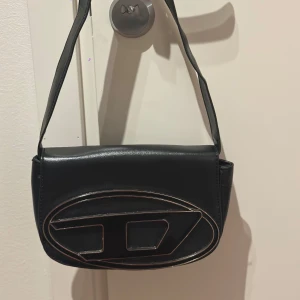 DIESEL axelväska - Svart axelväska i syntetmaterial med Diesel loggan på framsidan och stängs med en knapp. Köpt här på Plick men behövs inte längre så säljer den vidare. 