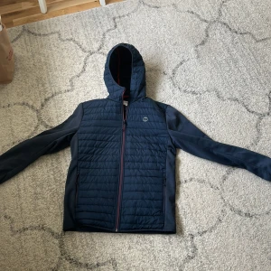 Mörkblå quiltad jacka - Säljer en mörkblå quiltad jacka med huva och dragkedja. Jackan har långa ärmar och en stilren design, perfekt för kyligare dagar. Den är både funktionell och snygg med en sportig look. Använd fåtal gånger så den är i perfekt skick.