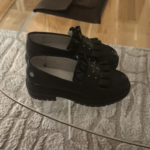 Svarta loafers med volangdetalj -  Svarta loafers med en volangdetalj och små nitar på ovansidan. Nyskick endast använda fåtal gånger i storlek 36. Nypris: 1400kr. 