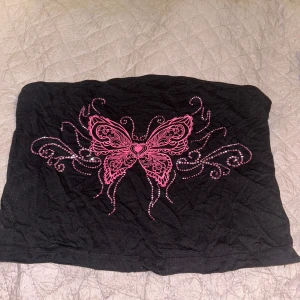 Svart tubtopp med rosa fjärilsmönster - Snygg svart tubtopp med ett stort rosa fjärilsmönster i mitten. Perfekt för en cool och avslappnad stil. Passar bra till jeans eller kjol för en trendig look.