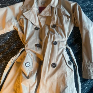 Beige trenchcoat - Snygg beige trenchcoat med klassisk design och knappar framtill. Jackan har ett bälte i midjan och långa ärmar. Perfekt för en stilren look. Det står dock inte storleken men skulle nog säga att det är en xxs 