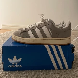 Grå Adidas Campus sneakers - Snygga grå Adidas Campus sneakers med vita detaljer och klassiska tre ränder. Skorna har en bekväm passform och är perfekta för en avslappnad stil. De är tillverkade i mocka och har snörning framtill.