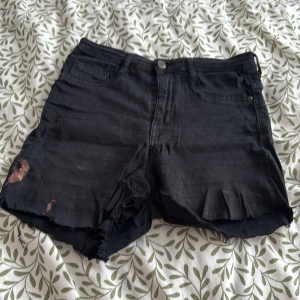 Svarta jeansshorts - Säljer ett par svarta jeansshorts med råa kanter och slitna detaljer. Shortsen har en knapp och dragkedja framtill samt klassiska fem fickor. Perfekta för en avslappnad stil.