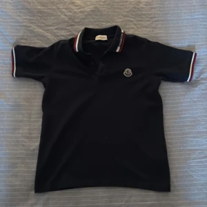 Mörkblå  piké från Moncler 140 - Säljer en stilren mörkblå pikétröja från Moncler med korta ärmar. Tröjan har en klassisk krage med röd, vit och blå randdetalj samt Monclers logotyp på bröstet. Perfekt för en avslappnad men snygg look.