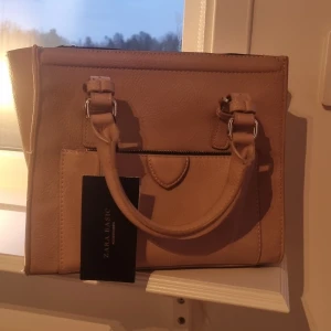 Beige handväska från Zara Basic - Snygg beige handväska från Zara Basic med praktiska dragkedjor och dubbla handtag. Perfekt för att hålla dina saker organiserade och lättillgängliga. Stilren design som passar till många tillfällen.