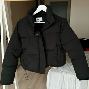 Svart pufferjacka från NA-KD - Säljer en svart pufferjacka från NA-KD. Jackan har en hög krage och stängs med dragkedja och knappar. Knappt använd. Perfekt för kyliga dagar med sin vadderade design 💓