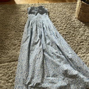 Blommig axelbandslös långklänning - Säljer en söt axelbandslös långklänning med blått blommönster. Klänningen har en smockad överdel som ger en bekväm passform och ett luftigt fall. Perfekt för sommarens varma dagar!