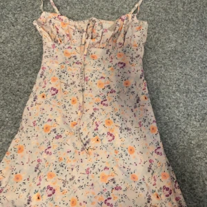 Blommig sommarklänning med smala axelband - Charmig blommig klänning med smala axelband och volangdetaljer vid bysten. Klänningen har ett färgglatt mönster med blommor i orange, lila och rosa nyanser på en ljus bakgrund. Perfekt för varma sommardagar.