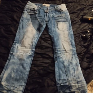 Blå jeansbyxor från Leashe - Snygga blå jeansbyxor från Leashe med unikt tvättat mönster och flera fickor. Byxorna har en cool design med detaljer som broderad text och kontrasterande sömmar. Perfekta för en avslappnad stil.