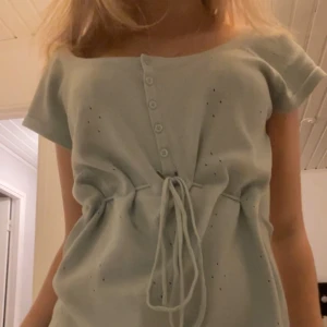 Ljusgrön topp med knyt och knappar - Säljer en söt ljusgrön topp med knappar framtill och knytband i midjan. Toppen har korta ärmar och ett diskret hålmönster som ger en fin detalj. Perfekt för en avslappnad stil.