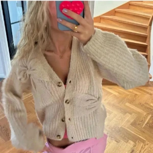 Stickad kofta - Mysig beige stickad kofta från Zara med knappar framtill och en dekorativ rosett på sidan. Perfekt för kyliga dagar med sin mjuka och varma känsla. Koftan har långa ärmar och en avslappnad passform. Den är köpt på barnavdelningen så storleken på lappen är 164 men den passar mig som är en S.