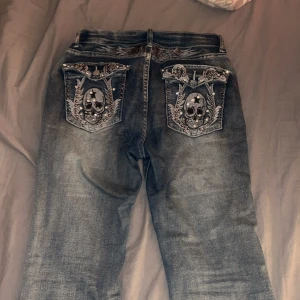Jeans med dödskallebroderi - Coola jeans med unikt dödskallebroderi på bakfickorna. De har en klassisk blå tvätt och en knappgylf. Perfekta för att ge din outfit en edgy touch.